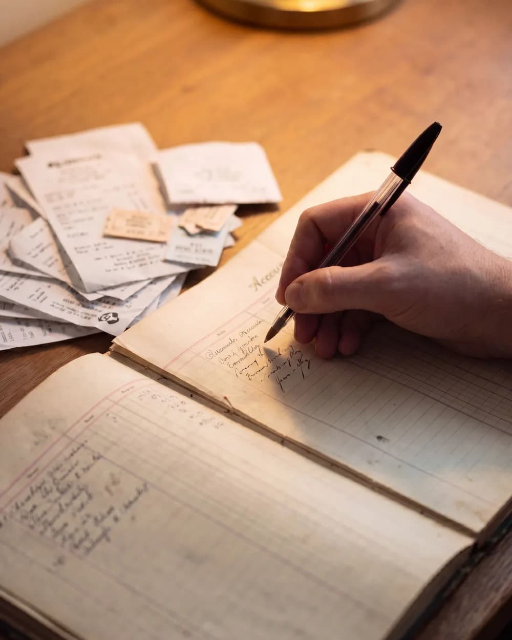 A hand writing an entry into an exhibitor accounts ledger next to a stack of receipts